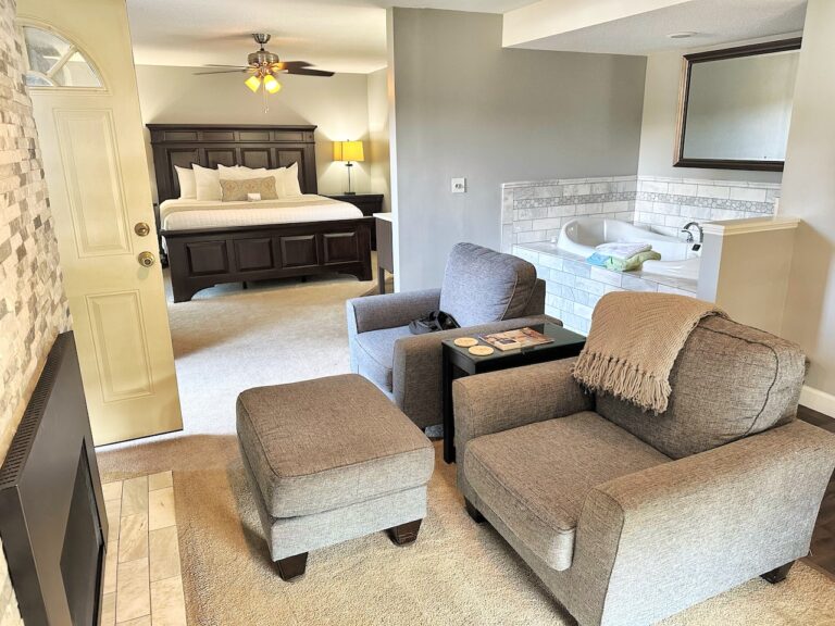 two gray lounge chairs with ottoman facing fireplace on left, large whirlpool tub behind chairs and king bed with dark wood foot and head board