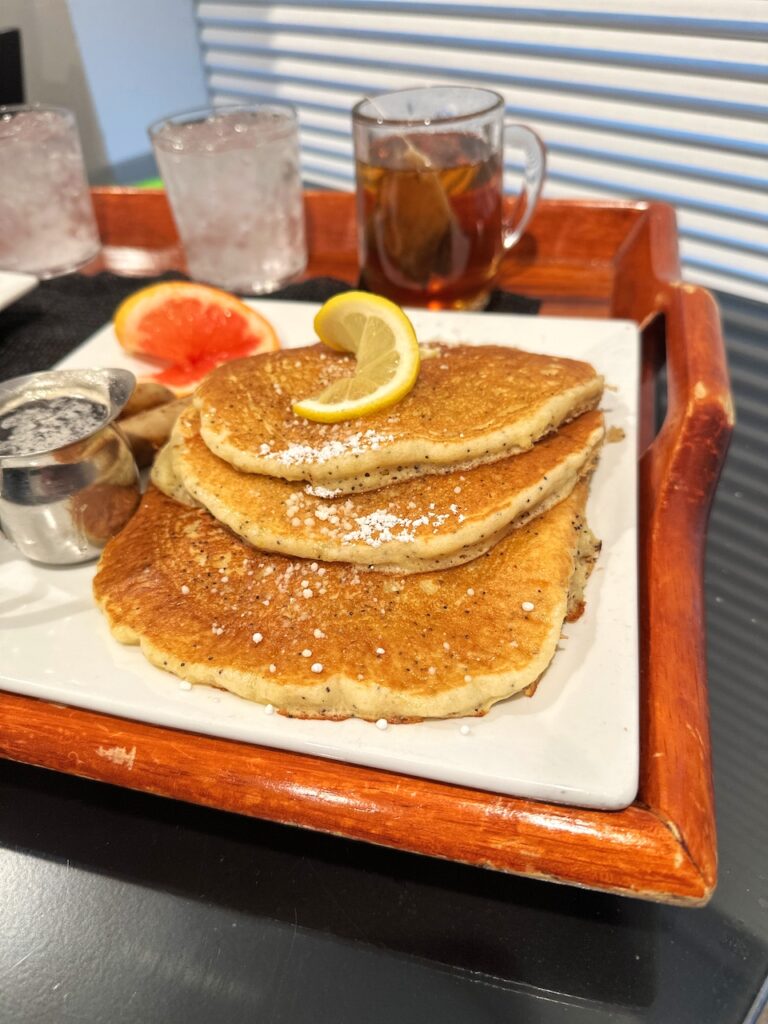 three fluffy pancakes with a lemon wedge on top and silver creamer cup of syrup on a white plate and a wood serving platter